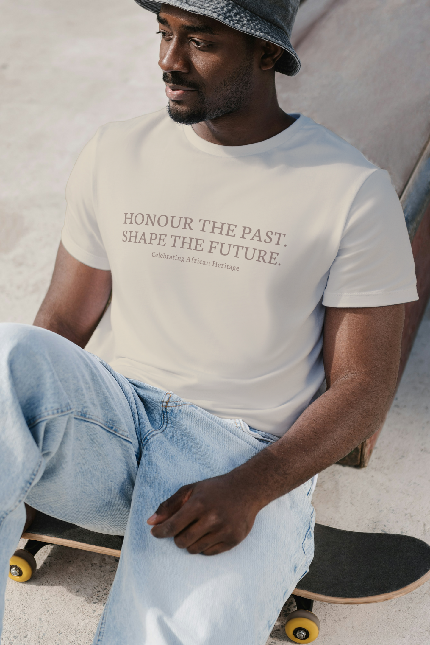 Model wearing a light-colored t-shirt with the text “Honour the Past. Shape the Future. Celebrating African Heritage", seated casually on a skateboard.