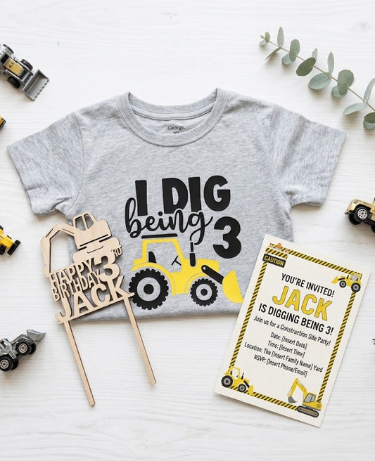 Flat lay of a construction-themed 3rd birthday party bundle including a grey "I Dig Being 3" t-shirt with a yellow excavator, a laser-cut wooden cake topper personalized for "Jack," and a matching "Construction Site Party" invitation. Part of The Birthday Edit Essentials Bundle.