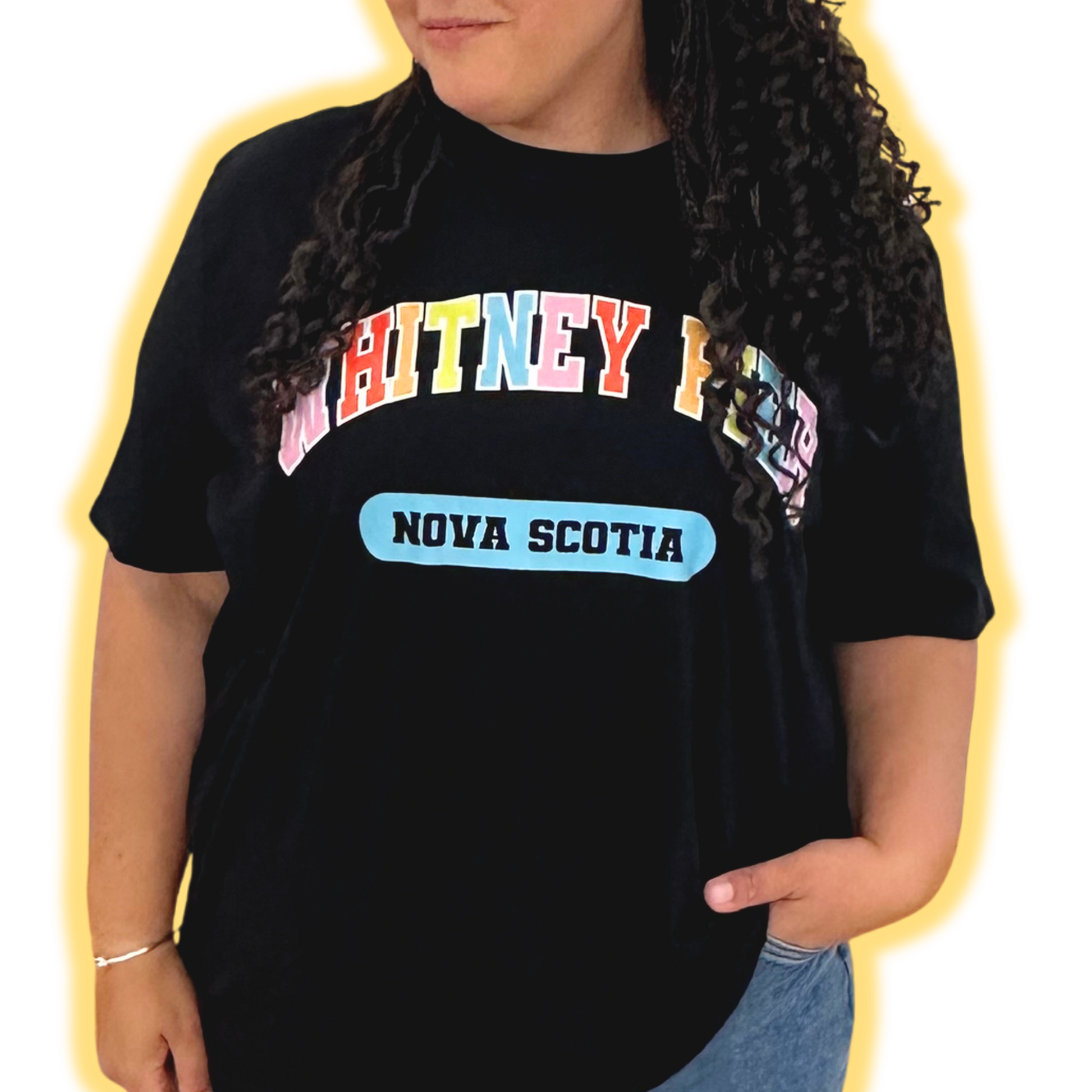 A woman wearing a black short-sleeve graphic t-shirt with 'WHITNEY PIER' in colourful block letters and a light blue 'NOVA SCOTIA' label on the front, set against a bright yellow background.