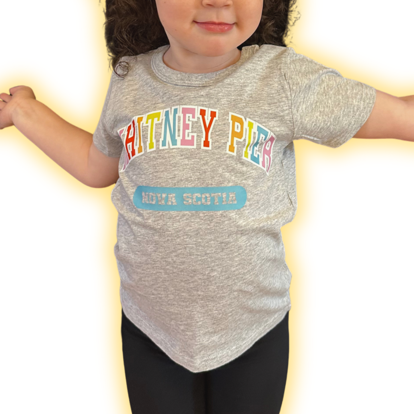 A toddler wearing a heather grey short-sleeve t-shirt with 'WHITNEY PIER' in colourful letters and a blue 'NOVA SCOTIA' patch graphic, paired with black leggings, set against a bright white background with a yellow glow effect.
